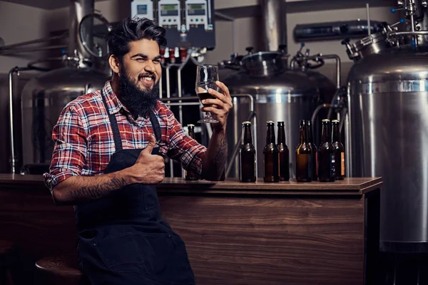 Şık tam Hintli bir yün gömlekli sakallı ve önlük bir bardak bira, bira fabrikası sayacında arkasında oturan tutar.