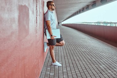 Portre yakışıklı bir moda Freelancer güneş gözlüğü beyaz bir gömlek giymiş ve şort uzağa arıyorsunuz köprünün altından bir duvara yaslanmış bir dizüstü bilgisayar ayakta tutan.