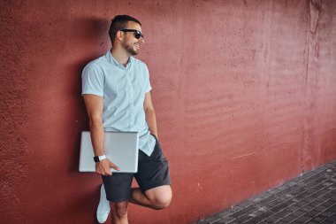 Beyaz bir gömlek ve şort giymiş güneş gözlüğü mutlu yakışıklı şık serbest meslek köprü altında bir duvara yaslanmış bir dizüstü bilgisayar ayakta tutan.