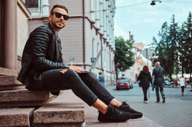 Moda serbest meslek adamı gülümseyen bir siyah ceket giymiş ve kot Avrupa'nın eski bir bina karşı adımlar üzerinde otururken dizüstü tutar.