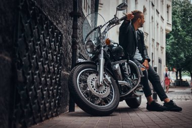 Bir siyah deri ceket ve retro motosikleti eski Avrupa sokakta oturan kot giymiş şık motorcu.