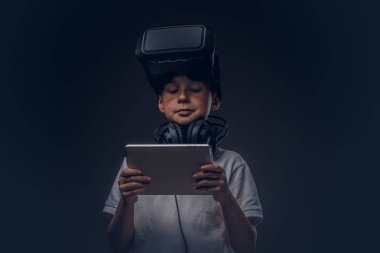 Küçük bir çocuk portresi olan sanal gerçeklik gözlük ve kulaklık beyaz bir tişört giymiş ve koyu arka plan üzerinde izole bir stüdyoya dijital tablet kullanma. Eğitim ve teknoloji kavramı.