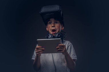 Sürpriz bir küçük çocuk portresi sanal gerçeklik gözlüklü beyaz tişört giymiş ve kulaklık dijital tablet koyu arka plan üzerinde izole bir stüdyoda tutarak. Eğitim ve teknoloji kavramı.
