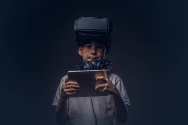 Beyaz tişört, sanal gerçeklik Gözlüklü Şirin küçük bir çocuk portresi giymiş ve kulaklık dijital tablet karanlık bir arka plan üzerinde izole bir stüdyoda tutarak. Eğitim ve teknoloji kavramı.