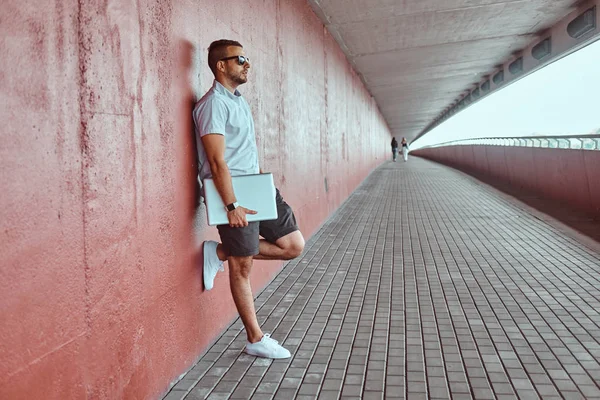 Portre yakışıklı bir moda Freelancer güneş gözlüğü beyaz bir gömlek giymiş ve şort uzağa arıyorsunuz köprünün altından bir duvara yaslanmış bir dizüstü bilgisayar ayakta tutan.