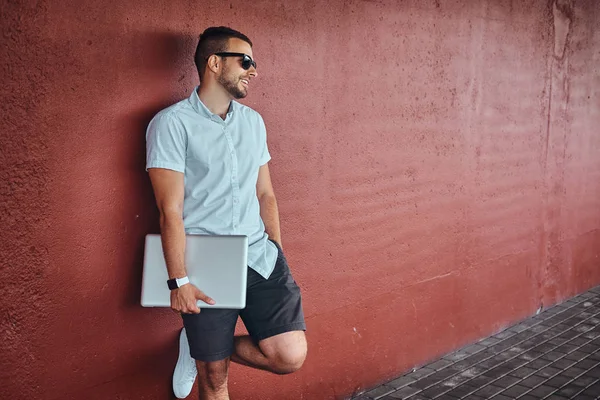 Beyaz bir gömlek ve şort giymiş güneş gözlüğü mutlu yakışıklı şık serbest meslek köprü altında bir duvara yaslanmış bir dizüstü bilgisayar ayakta tutan.