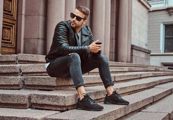 时髦的家伙穿着黑色夹克和牛仔裤持有智能手机坐在台阶上反对在欧洲的旧建筑 — 图库照片