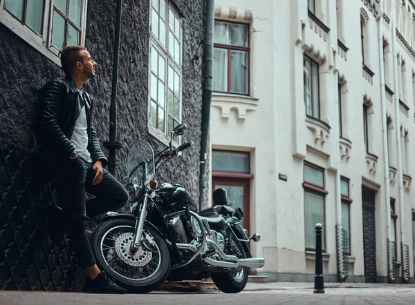 Bir siyah deri ceket ve kot eski bir Europe street retro motosikletine yakınındaki duvara yaslanmış şık motorcu giymiş.