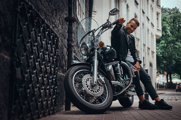 Happy fashionable biker dressed in a black leather jacket and jeans sitting on his retro motorcycle on old Europe street.