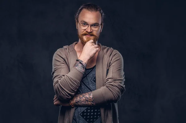 Smart pensive redhead hipster with full beard and glasses dressed in casual clothes, poses with hand on chin in a studio. Isolated on the dark background.
