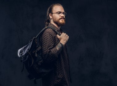 Gözlüklü bir kahverengi gömlek giymiş bir kızıl saçlı hipster öğrenci portresi tutan bir stüdyoda poz bir sırt çantası. Karanlık bir arka plan üzerinde izole. 