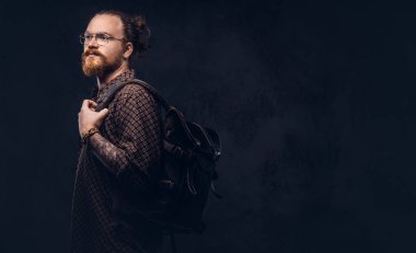 Gözlüklü bir kahverengi gömlek giymiş bir kızıl saçlı hipster öğrenci portresi tutan bir stüdyoda poz bir sırt çantası. Karanlık bir arka plan üzerinde izole. 