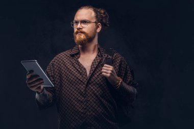 Gözlüklü bir kahverengi gömlek giymiş bir kızıl saçlı hipster öğrenci portresi tutan bir sırt çantası ve bir stüdyoda poz dijital tablet. Koyu arka plan üzerinde izole.