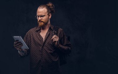 Gözlüklü bir kahverengi gömlek giymiş bir kızıl saçlı hipster öğrenci portresi tutan bir sırt çantası ve bir stüdyoda poz dijital tablet. Koyu arka plan üzerinde izole.