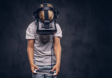 Sarı saçlı Genç çocuk sanal gerçeklik gözlük takan ve dijital tablet kullanma beyaz bir t-shirt giymiş. Karanlık dokulu bir arka plan üzerinde izole.