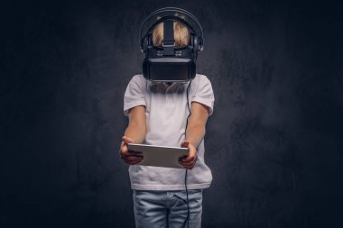 Liseli Sarışın sanal gerçeklik gözlük takan ve dijital tablet kullanma beyaz bir t-shirt giymiş. Karanlık dokulu bir arka plan üzerinde izole.