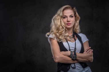 Kıvırcık sarışın kız sevimli beyaz bir t-shirt giymiş ve aksesuarları ve kol saati, bir stüdyoda çapraz silah ile poz bir sürü yelek giyer. Karanlık dokulu bir arka plan üzerinde izole.