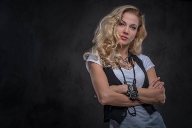 Şehvetli Kıvırcık sarışın kadın beyaz bir t-shirt giymiş ve aksesuarları ve kol saati, bir stüdyoda çapraz silah ile poz bir sürü yelek giyer. Karanlık dokulu bir arka plan üzerinde izole.