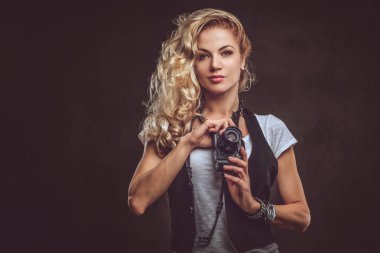 Şehvetli Kıvırcık sarışın kız fotoğrafçı beyaz bir t-shirt giymiş ve aksesuarları ve kol saati, bir stüdyoda bir kamera ile poz bir sürü yelek giyer. Karanlık dokulu arka plan üzerinde izole.
