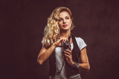 Güzel Kıvırcık sarışın kadın fotoğrafçı beyaz bir t-shirt giymiş ve aksesuarları ve kol saati, bir stüdyoda bir kamera ile poz bir sürü yelek giyer. Karanlık dokulu arka plan üzerinde izole.