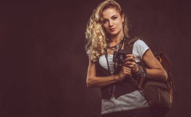 Sevimli Kıvırcık sarışın kadın giyinmiş bir beyaz gömlek ve yelek giyer aksesuarları ve kol saati, bir sürü bir kamera ve karanlık dokulu bir arka plan üzerinde izole bir stüdyoda sırt çantası ile poz. Macera ve Turizm kavramı.