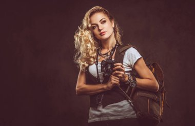 Şehvetli Kıvırcık sarışın kadın giyinmiş bir beyaz gömlek ve yelek giyer aksesuarları ve kol saati, bir sürü bir kamera ve karanlık dokulu bir arka plan üzerinde izole bir stüdyoda sırt çantası ile poz. Macera ve Turizm kavramı.