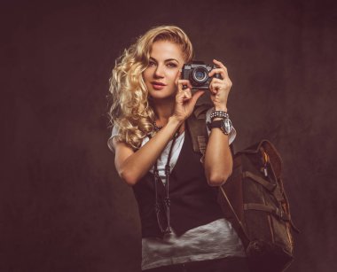 Şehvetli Kıvırcık sarışın kadın giyinmiş bir beyaz gömlek ve yelek giyer aksesuarları ve kol saati, bir sürü bir kamera ve karanlık dokulu bir arka plan üzerinde izole bir stüdyoda sırt çantası ile poz. Macera ve Turizm kavramı.