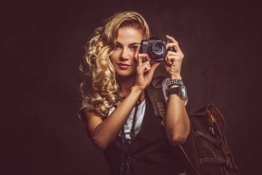 Sevimli Kıvırcık sarışın kadın giyinmiş bir beyaz gömlek ve yelek giyer aksesuarları ve kol saati, bir sürü bir kamera ve karanlık dokulu bir arka plan üzerinde izole bir stüdyoda sırt çantası ile poz. Macera ve Turizm kavramı.