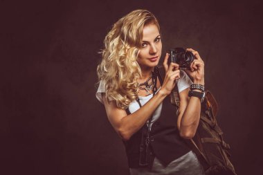 Sevimli Kıvırcık sarışın kadın giyinmiş bir beyaz gömlek ve yelek giyer aksesuarları ve kol saati, bir sürü bir kamera ve karanlık dokulu bir arka plan üzerinde izole bir stüdyoda sırt çantası ile poz. Macera ve Turizm kavramı.