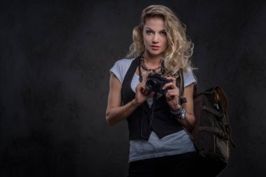 Sevimli Kıvırcık sarışın kadın giyinmiş bir beyaz gömlek ve yelek giyer aksesuarları ve kol saati, bir sürü bir kamera ve karanlık dokulu bir arka plan üzerinde izole bir stüdyoda sırt çantası ile poz. Macera ve Turizm kavramı.