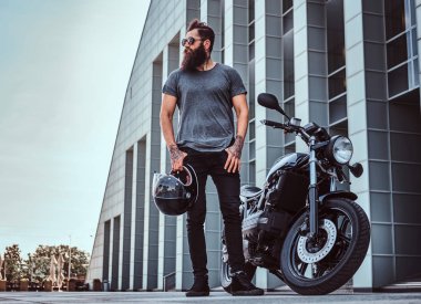 Gri tişört ve siyah pantolon yakınındaki özel yapım retro motosiklet bir gökdelen karşı ayakta acımasız sakallı erkek güneş gözlüğü giymiş.