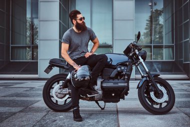 Acımasız sakallı erkek gri tişört ve siyah pantolon bir gökdelen karşı özel yapım retro motosiklet üzerinde oturan bir kask tutar.