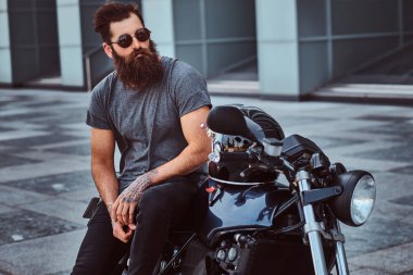 Gri tişört ve siyah pantolon bir gökdelen karşı özel yapım retro motosiklet üzerinde oturan bir acımasız sakallı bisikletçinin güneş gözlüğü portresi giymiş