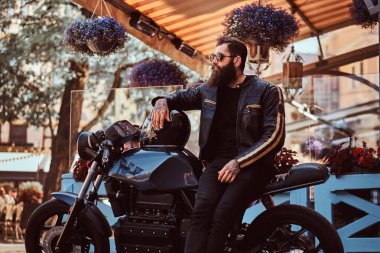 Güneş gözlüklü bir kafe Teras yakınındaki özel yapım retro motosiklet üzerinde oturan bir siyah deri ceket giymiş şık şık motorcu.