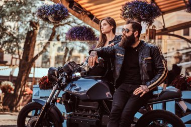 Çekici hipster çift - sakallı bir kafe Teras karşı poz vahşi erkek güneş gözlüğü giymiş bir siyah deri ceket ve onun genç seksi kız yanında, ayakta.