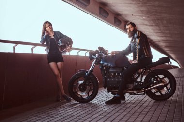 Portre çekici bir çift - vahşi motorcu bir motosiklet ve bir kameraya bakarak Genç Seksi esmer kız arkadaşına bir footway bir köprü altında oturan siyah deri ceketli sakallı.