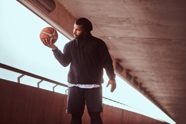 Köprü altında bir footway üzerinde bir basketbol duran siyah bir hoodie ve spor şort giymiş esmer bir adam portresi tutar.