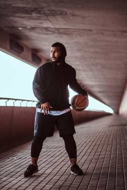 Esmer adam portresi köprü altında bir footway basketbol oynamak siyah bir hoodie ve spor şort giymiş.