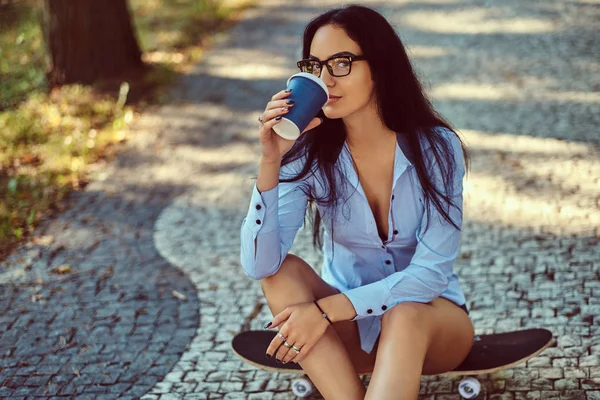 戴眼镜的性感黑发女孩穿着衬衫和短裤坐在滑板上 在公园里喝咖啡 — 图库照片