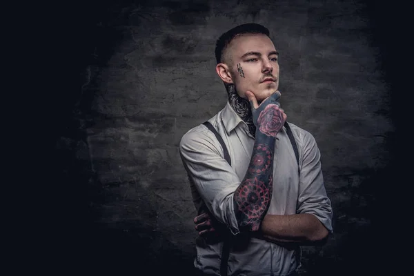 Close-up portrait of a thoughtful young old-fashioned tattooed guy wearing white shirt and suspenders holds hand on chin. Isolated on a dark background.