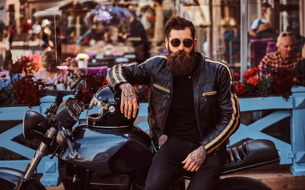 カフェのテラスの近く彼のカスタムメイドのレトロなバイクに座っている黒革のジャケットを着たサングラスのスタイリッシュなおしゃれなバイカー — ストック写真
