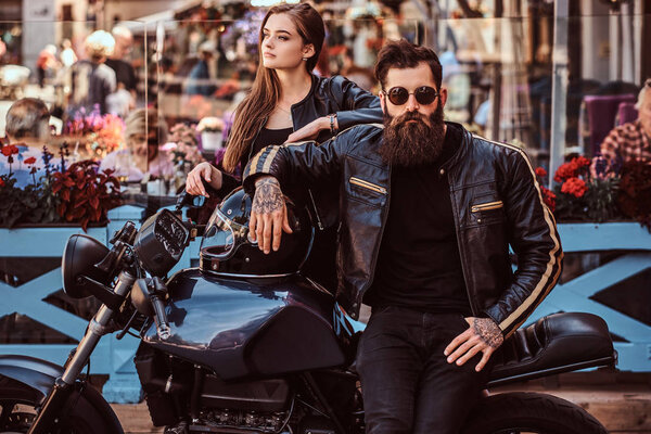 Attractive hipster couple - bearded brutal male in sunglasses dressed in a black leather jacket and his young sensual girl standing near, posing against terrace of a cafe.