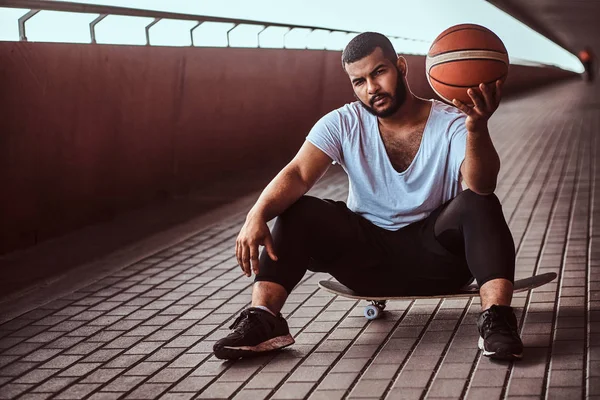 Pensive dark-skinned guy dressed in a white shirt and sports shorts holds a basketball while sitting on a skateboard on a footway under a bridge, looking at a camera.