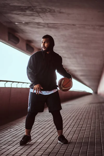 Portrait of a dark-skinned guy dressed in a black hoodie and sports shorts playing basketball on a footway under a bridge.