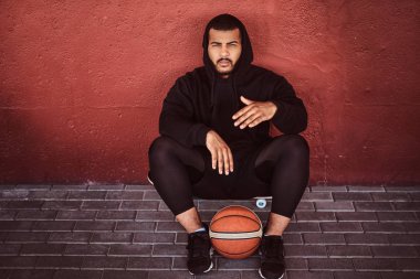 Afrikalı-Amerikalı basketbol ile bir kaykay üzerinde oturan ve duvara yaslanmış bir siyah hoodie ve spor şort giymiş adam sakallı.