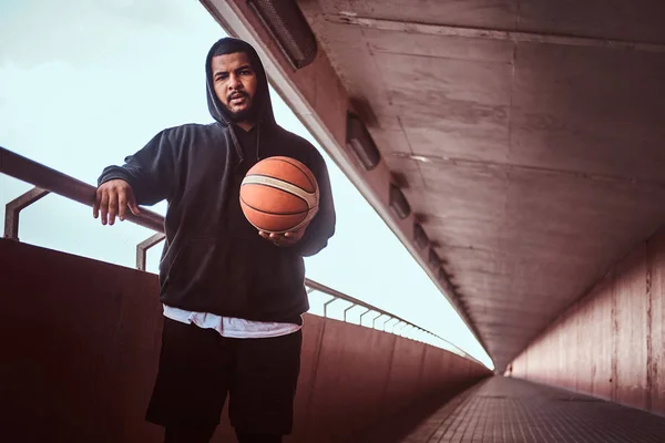 Portrait of a dark-skinned bearded guy dressed in a black hoodie and sports shorts leaning on a guardrail while standing with basketball.