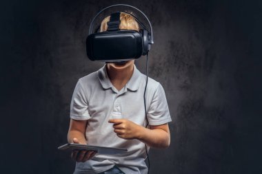 Sarı saçlı Genç çocuk sanal gerçeklik gözlük takan ve dijital tablet kullanma beyaz bir t-shirt giymiş. Karanlık dokulu bir arka plan üzerinde izole.