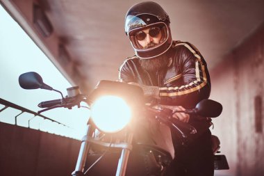 Acımasız bir sakallı motosiklet kask ve güneş gözlüğü yakın çekim portre dahil Far ile retro bir motosiklet üzerinde oturan bir siyah deri ceket giymiş.
