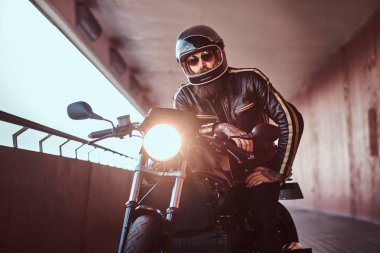 Acımasız bir sakallı motosiklet kask ve güneş gözlüğü yakın çekim portre dahil Far ile retro bir motosiklet üzerinde oturan bir siyah deri ceket giymiş.