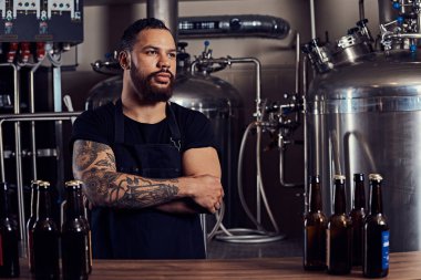 Portrait of a stylish bearded tattooed dark skinned male with crossed arms standing behind the counter in the brewery. clipart
